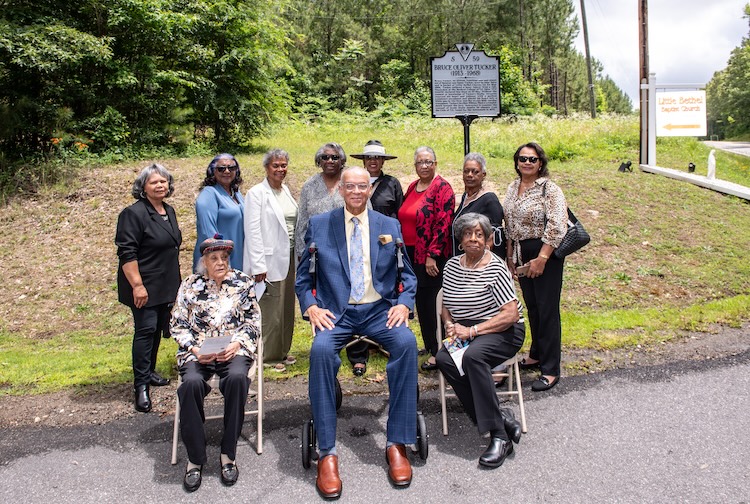 브루스 터커의 가족들이 새로운 역사적 고속도로 표시 앞에 앉아서 서 있습니다.
