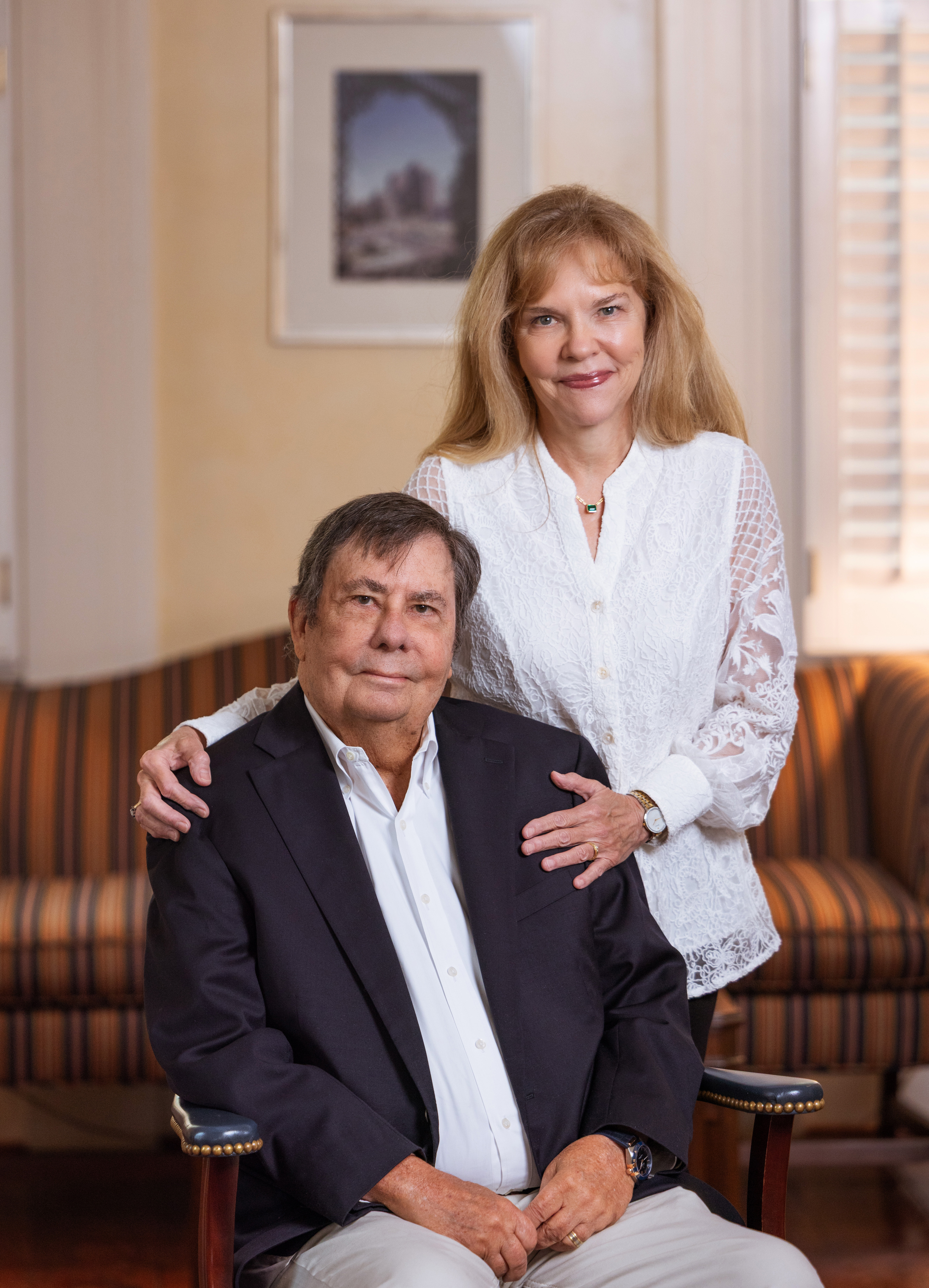 정장을 입고 앉아 있는 남자와 그 뒤에 흰 블라우스를 입은 여자가 서 있습니다.