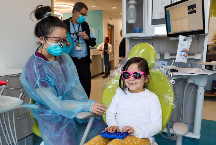 선글라스를 쓴 어린 소녀가 의자에 앉아있습니다. 학생이 옆에 서서 약속을 준비하고 있습니다.