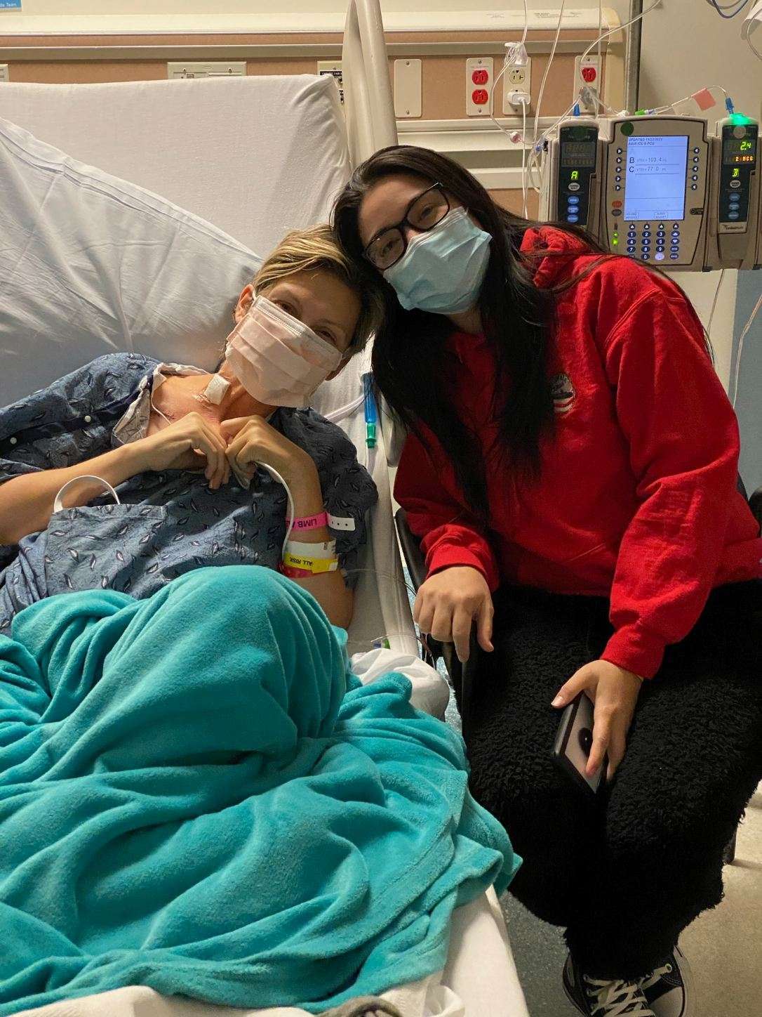 병실에 두 명의 여성이 있습니다. 한 여성은 침대에 앉아 있고 다른 여성은 그녀 옆에 서 있습니다.