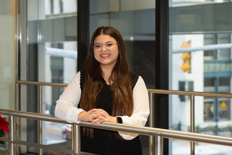 여성의 상반신 사진입니다. 그녀는 미소를 지으며 앞에 있는 금속 난간에 양손을 얹고 있습니다.